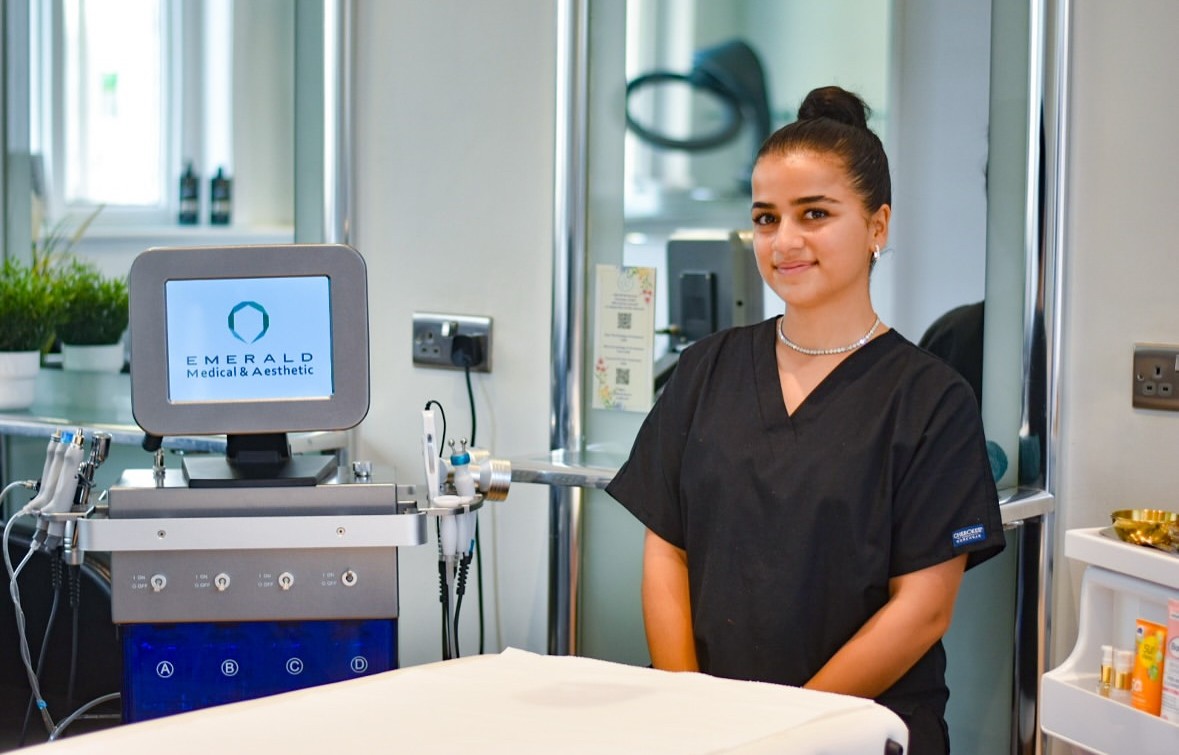 Smiling medical professional in black scrubs standing next to a medical aesthetic machine in a clean treatment room.