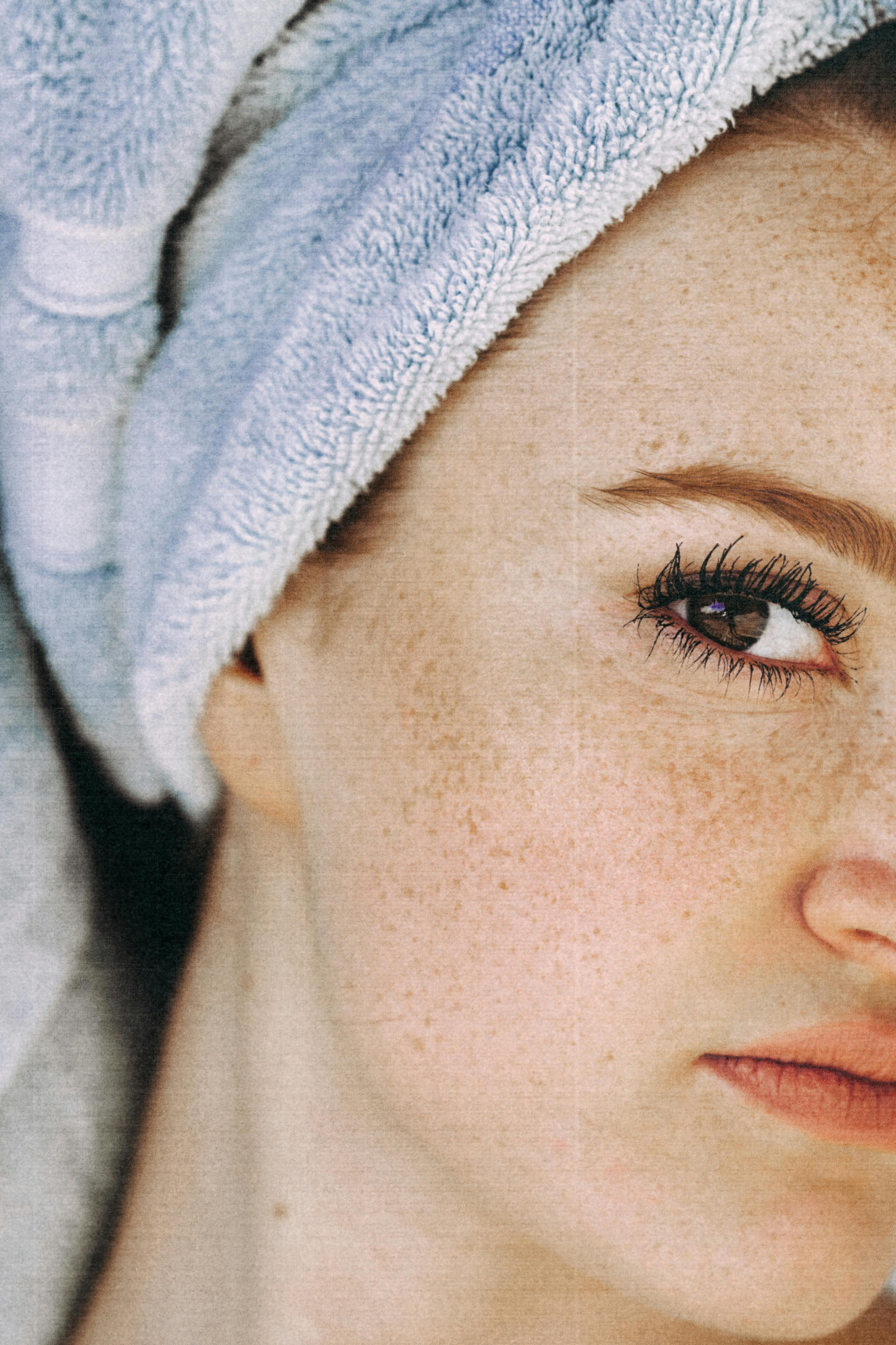 Close-up of a woman's freckled face with long eyelashes and a light blue towel wrapped around her head.