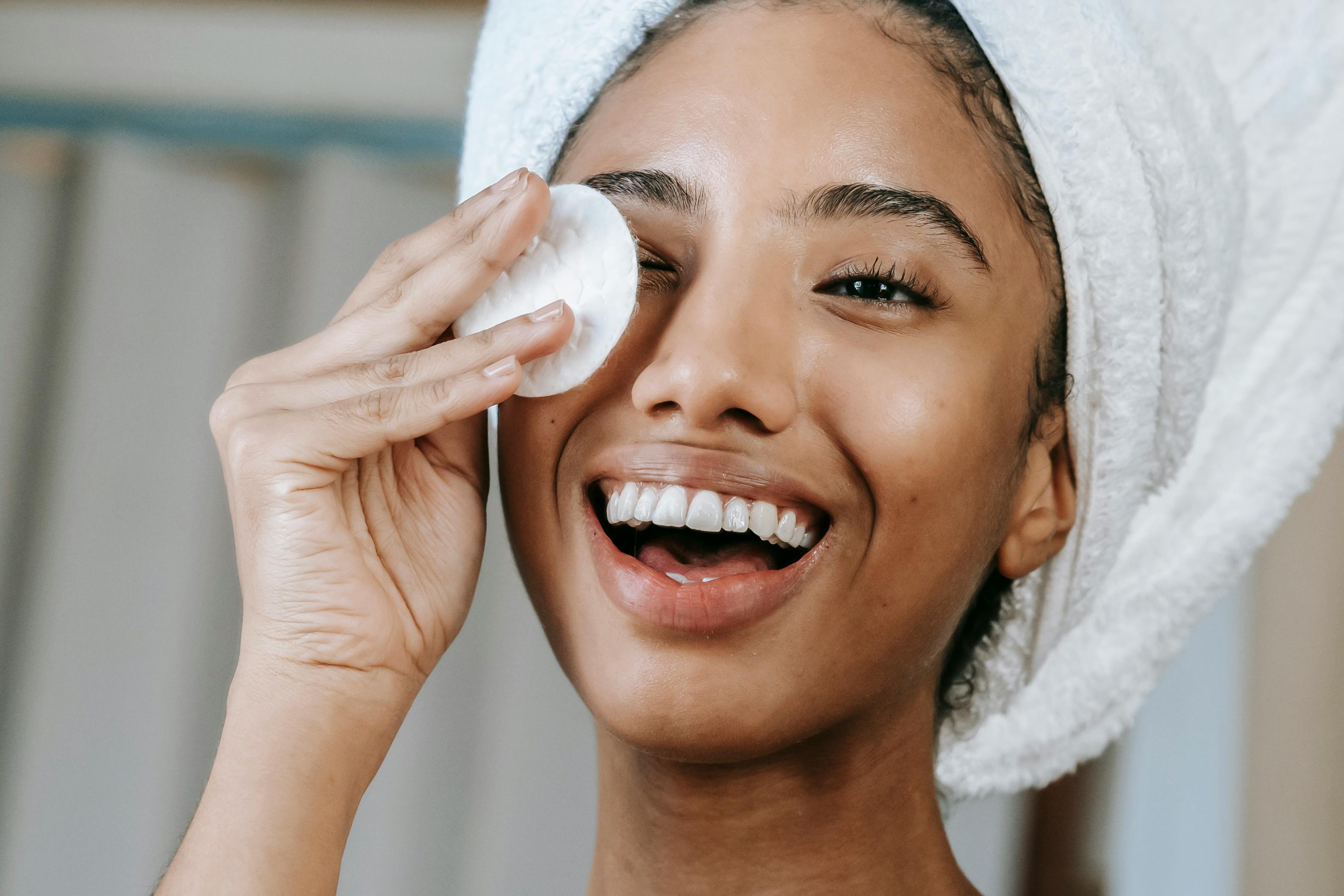 Smiling woman with a white towel wrapped around her head cleansing her face with a cotton pad.